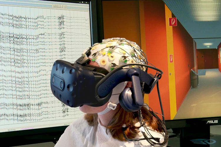 Person mit aufgesetzter Virtual-Reality-Brille und EEG. Im Hintergrund sind EEG-Daten und die virtuelle Umgebung zu sehen.