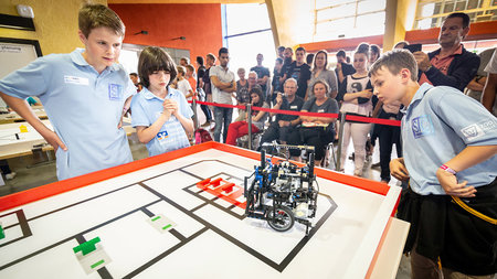 Drei Schüler beobachten, die ein Roboter über den Wettkampf-Tisch fährt.