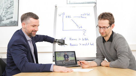 Zwei Männer sitzen vor einem Tablet Computer. Ein Mann zeigt mit seiner bionischen Handprothese an eine Tafel.