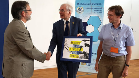 Ein älterer Mann nimmt ein gerahmtes Bild von einem alten Mann entgegen. Darauf steht: 58. Bundesrunde Mathematik-Olympiade 2019 in Chemnitz.  