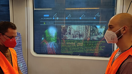 Zwei Männer mit Grsichtsmaske sitzen in einem Zugabteil, einer schreibt an einem Laptop. Am Zugfenster ist eine Projektion zu erkennen.