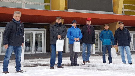 Sechs ältere Männer in winterlicher Kleidung stehen vor einem Gebäude im Schnee.