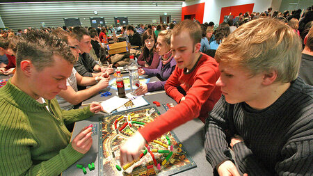 Viele junge Menschen sitzen in einem großen Raum an Tischen und spielen miteinander.