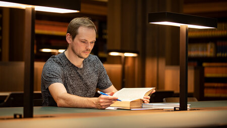Ein Mann sitzt an einem Tisch in einem Lesesaal und blickt in ein Buch.
