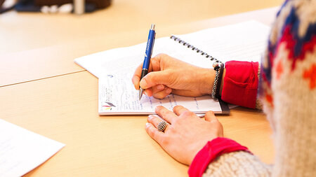 Eine an einem Tisch sitzende Person schreibt etwas auf ein Blatt Papier. 