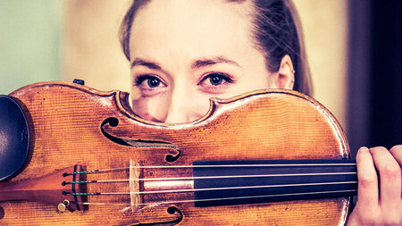 Eine junge Frau blickt ber ihre Violine, die sie quer vor ihr Gesicht hlt.