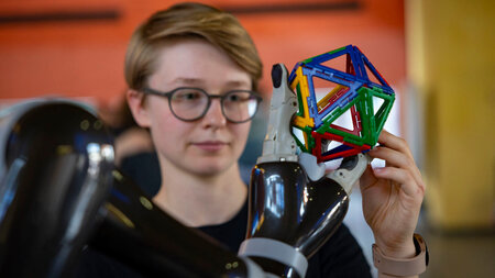 Eine junge Frau blickt auf eine Roboterhand, die ein buntes geometrisches Gebilde festhlt.