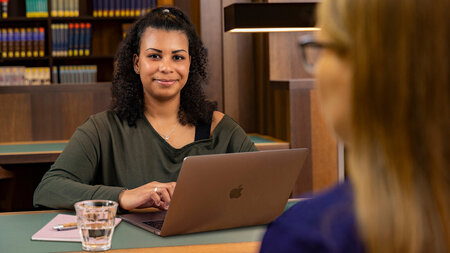 Eine Person sitzt in einem Lesesaal an einem Tisch vor einem Laptop und blickt Eine Frau，die gegenüber sitzt-an。