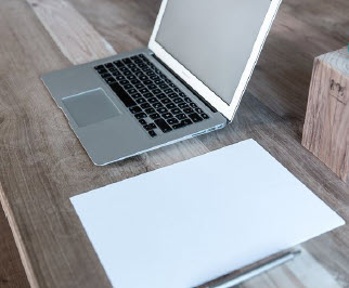 Ein Laptop steht auf dem Tisch und ein Blatt Papier liegt mit einem Stift bereit.