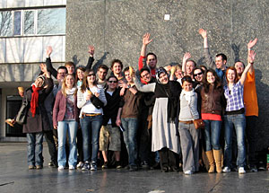 Bild mit großer Gruppe internationaler Studierender vor einem grauen Gebäude, die die Hände in die Luft halten.