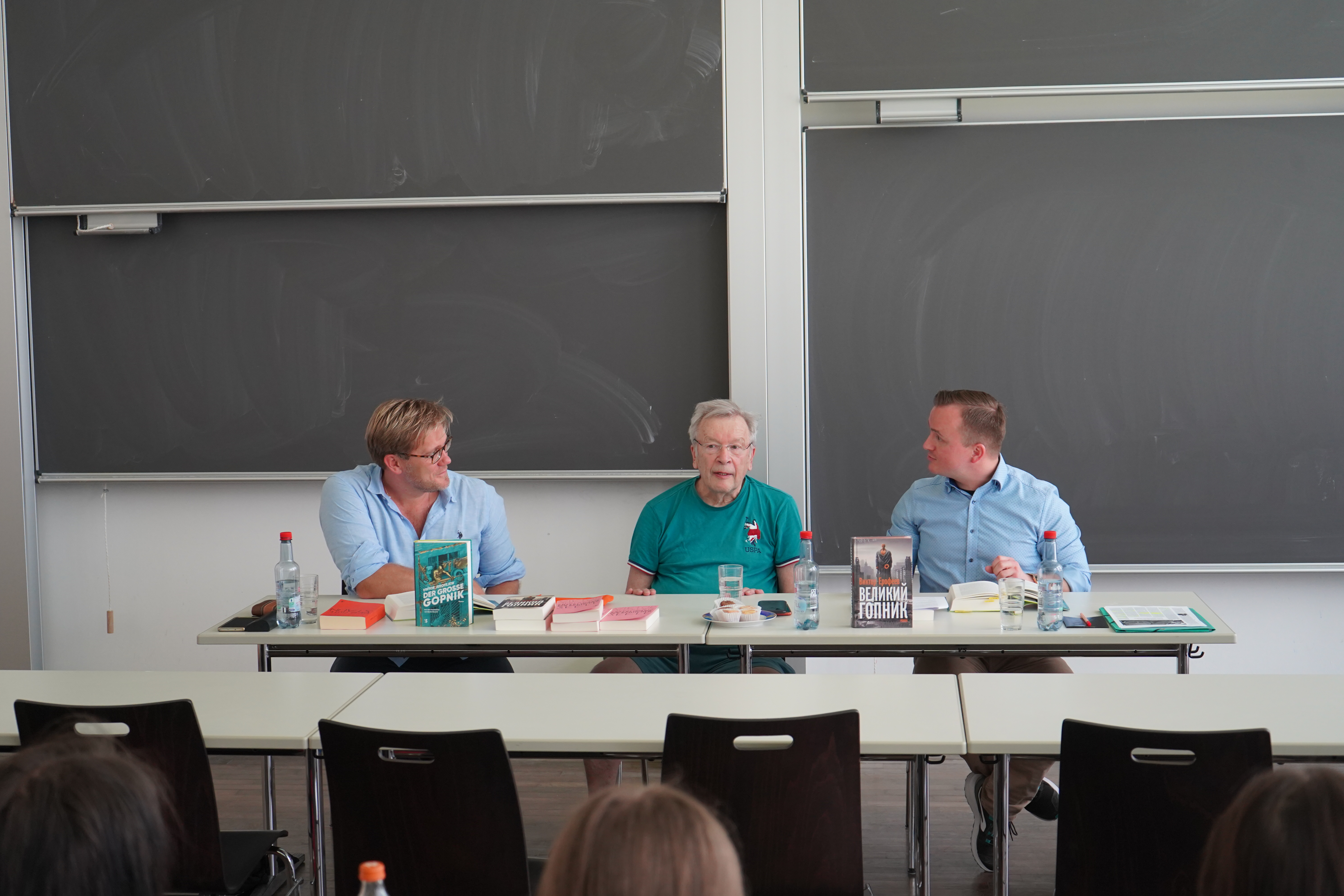 Der russische Schriftsteller und Dissident Viktor Jerofejew vor Studierenden der Seminare: „Russland unter Putin“ und „Alexander Kluge“, moderiert von Doktor Eugen Wenzel, Neuere Deutsche und Vergleichende Literaturwissenschaft (links) und Doktor Jakob Kullik, Internationale Politik (rechts); hinter Ihnen an der Wand eine grüne Tafel; auf dem Tisch sind zwei Bücher von Viktor Jerofejew aufgestellt