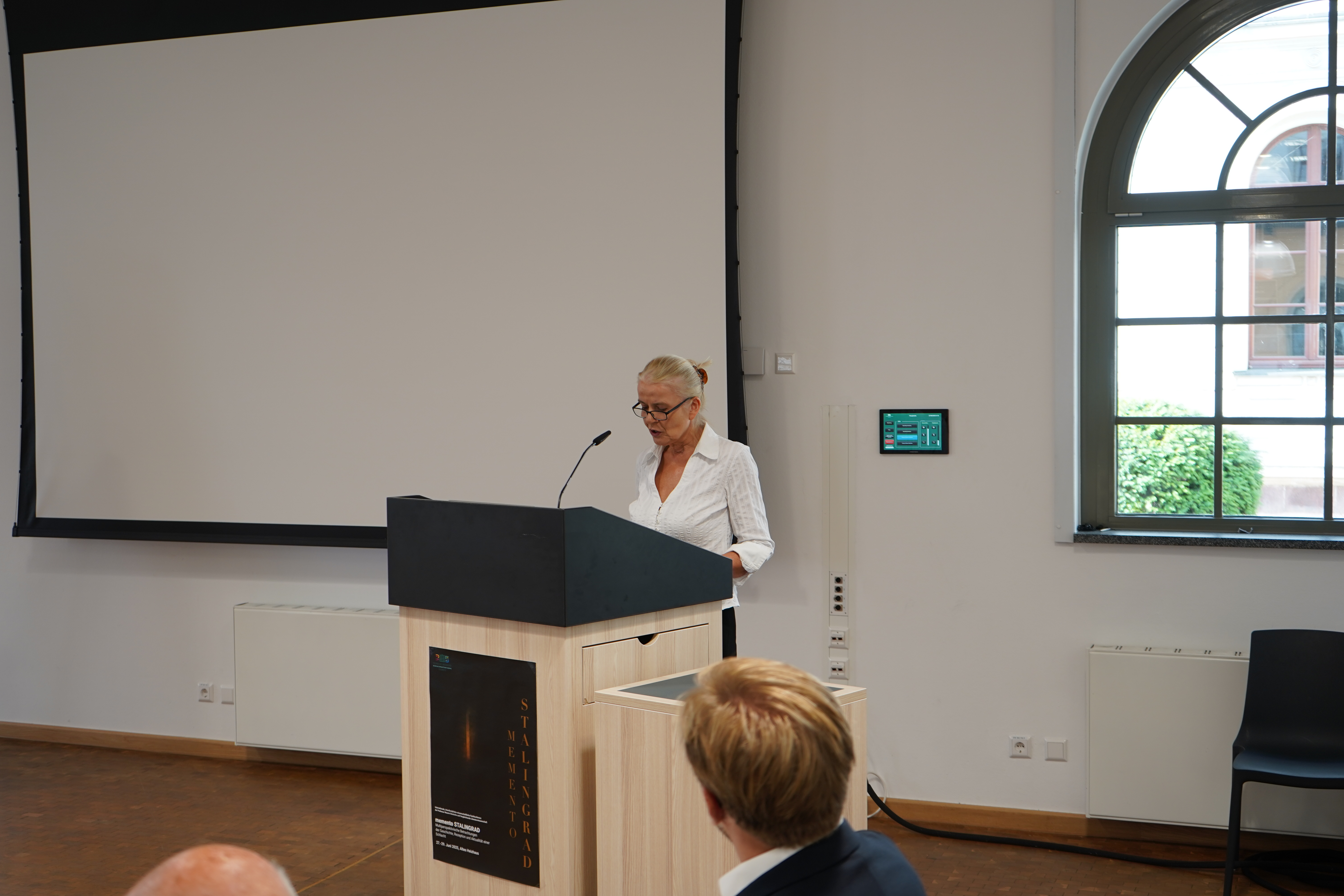 Professorin Bernadette Malinowski eröffnet offiziell die Konferenz; stehend hinter einem Pult, vor einer weißen Wand mit Leinwand