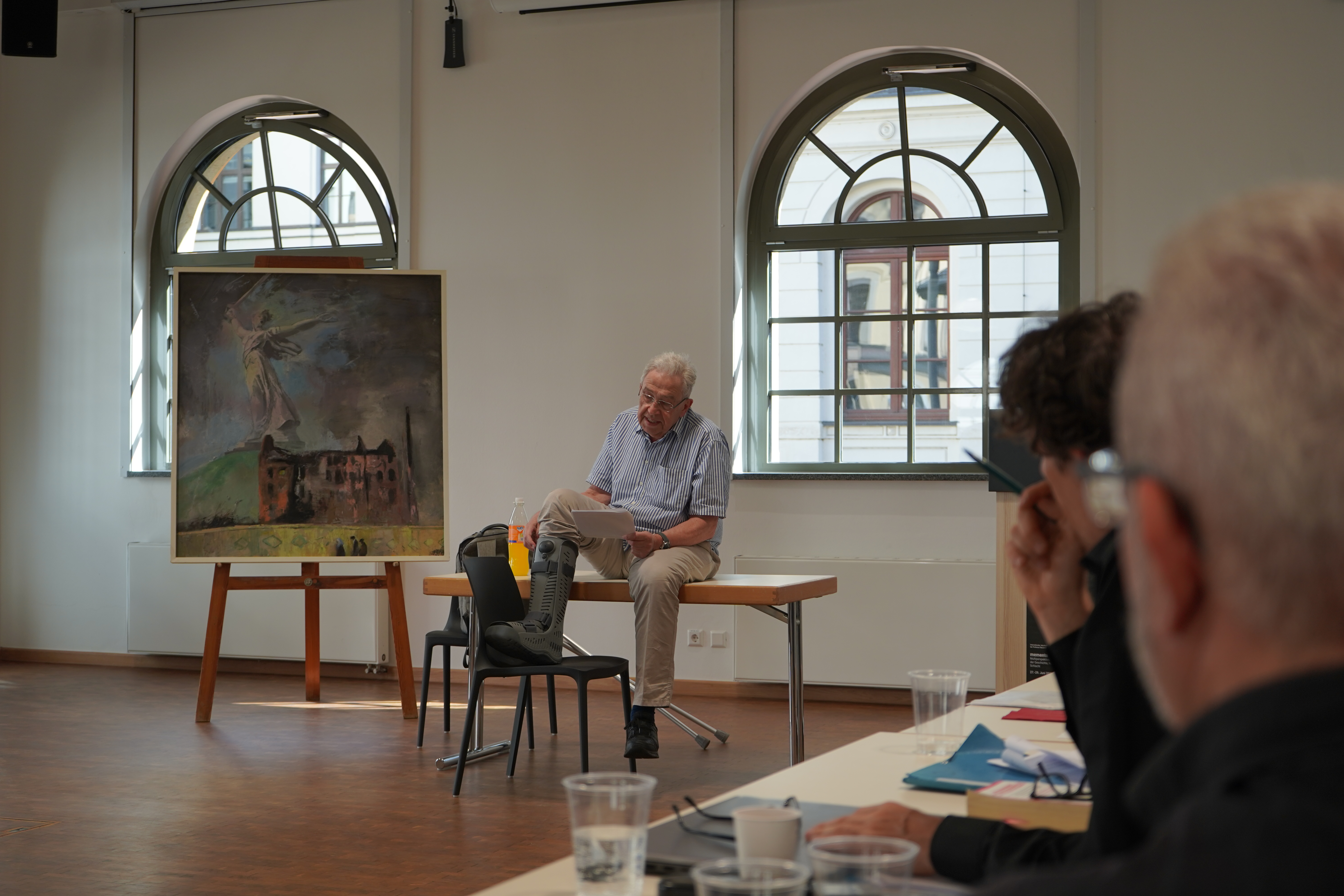Trotz körperlicher Beeinträchtigung referiert Prof. Dr. Hans-Ludwig Kröber zu Stalingrad aus dem Blickwinkel der forensischen Psychiatrie; sitzend auf einem Tisch, vor sich in der Hand hält er Zettel und hat sein geschientes Bein auf einem Stuhl abgesetzt