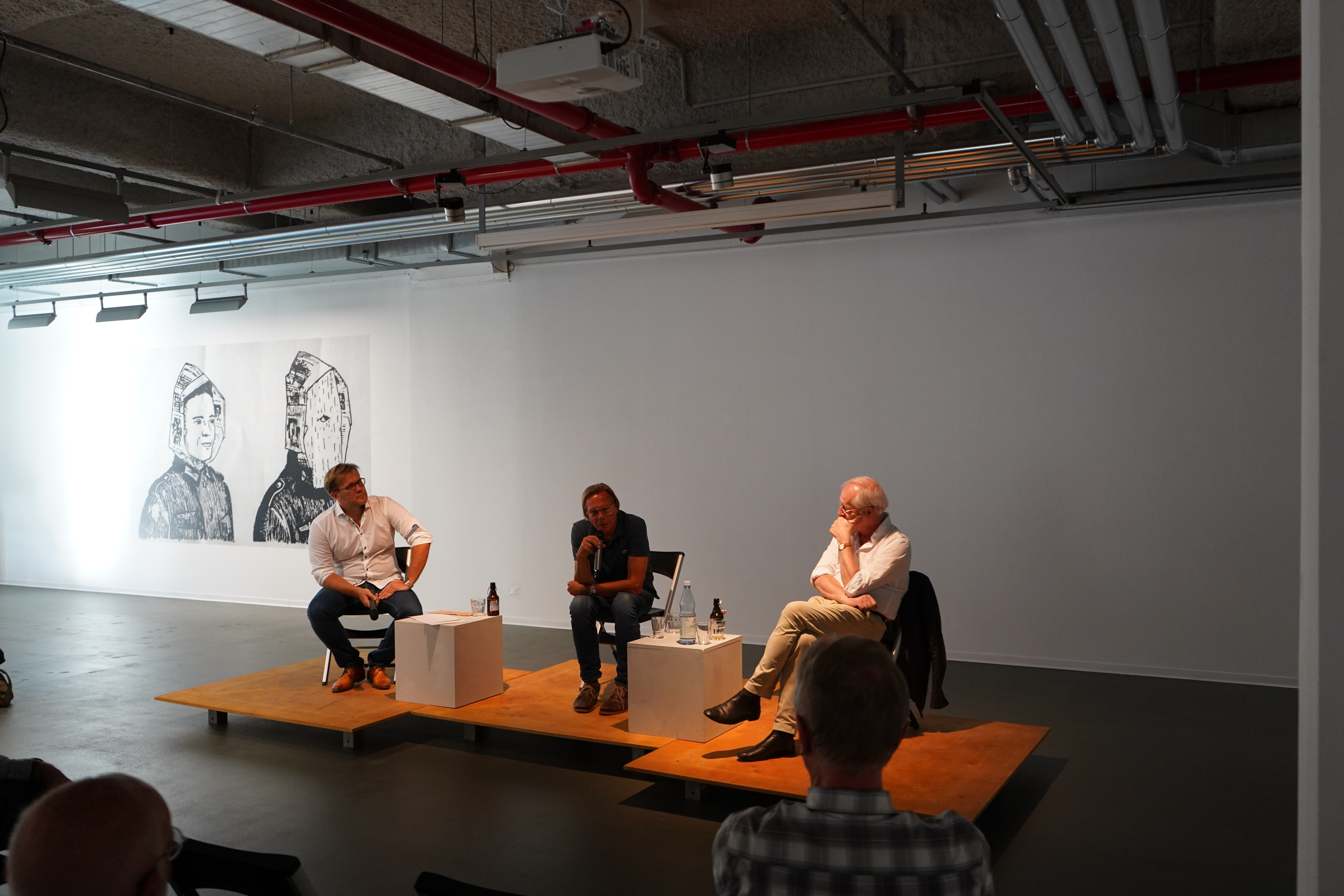 Professor Harald Welzer (in der Mitte des Bilde) und Professor Jörg Baberowski (rechts im Bild) in der Diskussion, moderiert von Dr. Eugen Wenzel (linksim Bild); das Podium steht leicht erhöht auf Holzplatten, zwischen den sitzenden Teilnehmern befinden sich kleine Tische mit Getränken, im Hintergrund eine weiße Wand, auf der sich auf der linken Seite eine Bild mit zwei Personen befindet