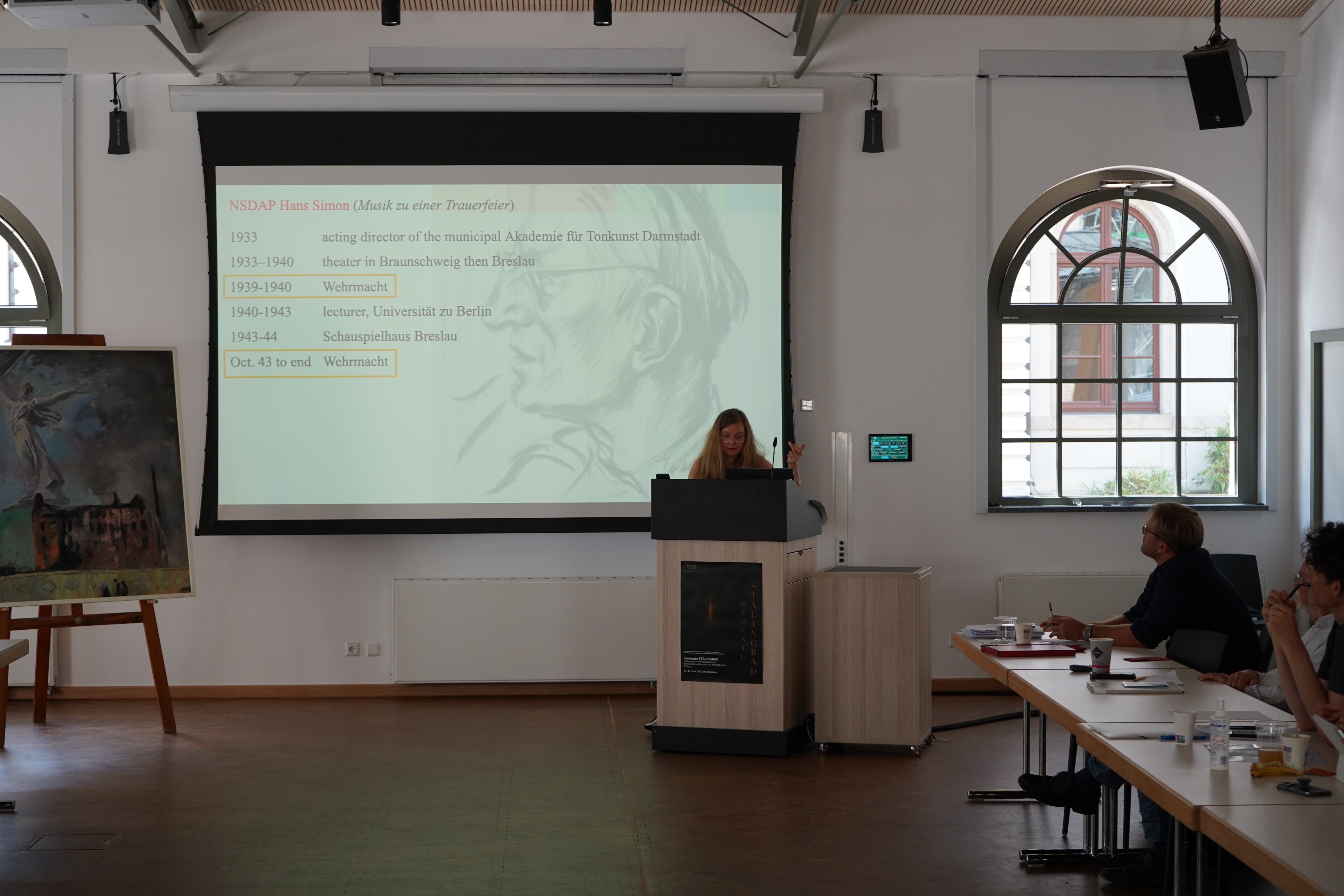  Professorin Karen Painter spricht über die deutsche Musikkomposition nach der Schlacht um Stalingrad; im Hintergrund befindet sich eine Leinwand, die auf einer Präsentationsfolie Jahresdaten und Angaben der Tätigkeit von Hans Simon (NSDAP) zeigt