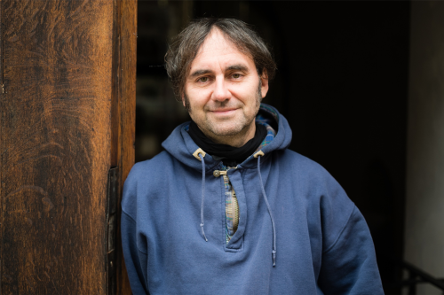 Das Foto zeigt den Autor Geralf Pochop im Jahr 2017. Er lehnt in einer Tür mit Holzrahmen, trägt einen blauen Kapuzenpullover und lächelt freundlich in die Kamera. 