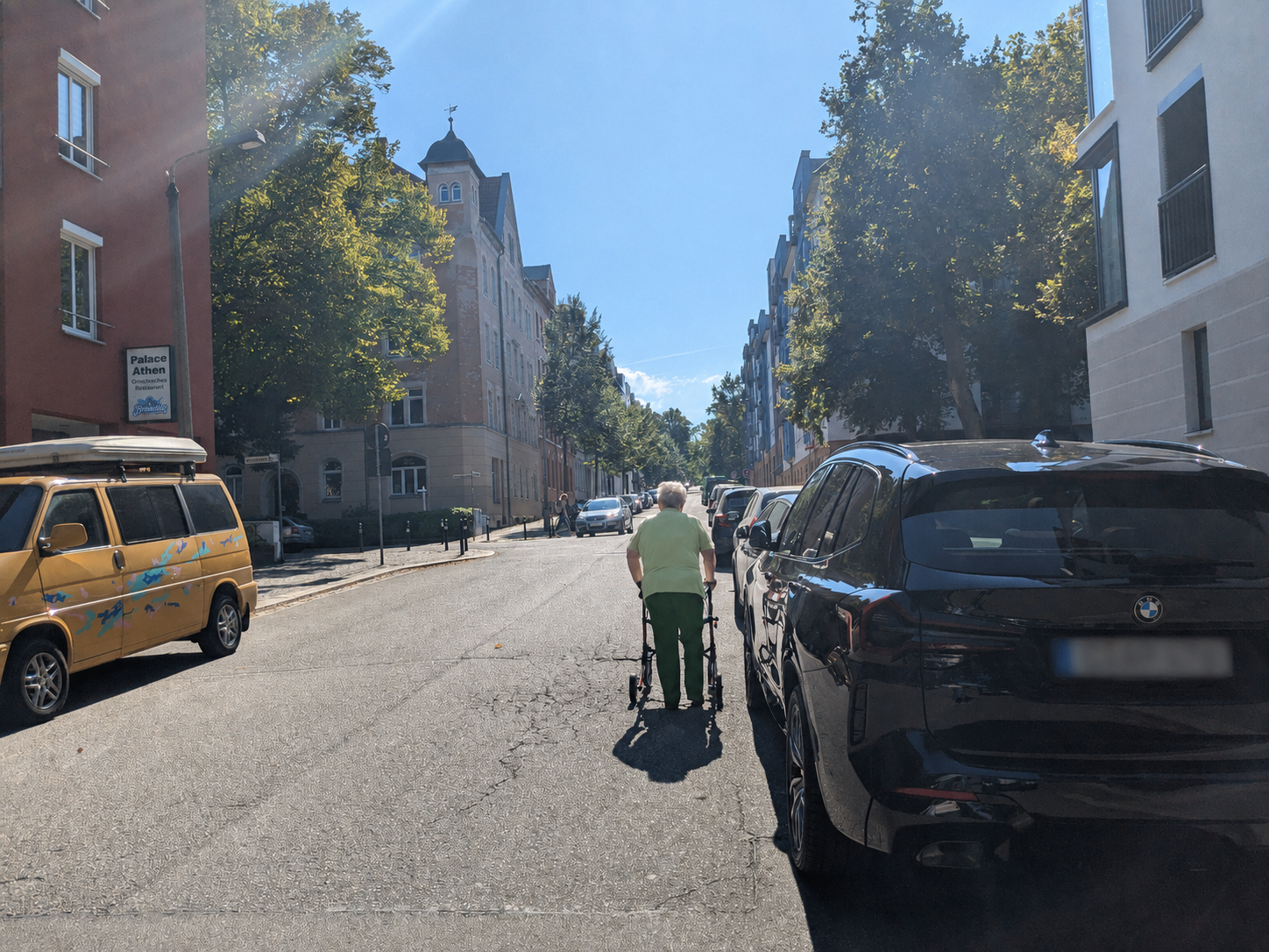 Eine Person im Rollstuhl fährt mit einer Begleitperson auf der Fahrbahn einer herbstlichen Wohnstraße. Beide Straßenseiten sind dicht mit Autos beparkt; auf den Gehwegen liegen viele Blätter. Mehrstöckige Wohnhäuser und gelb-orange gefärbte Bäume säumen die Straße.