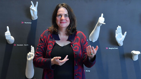 Frau gestikuliert, während an der Wand im Hintergrund künstliche Hände verschiedene Gesten zeigen