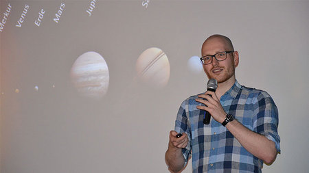 Mann mit Brille und blauem Hemd hält eine Rede per Mikrofon