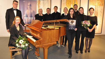 Eine Frau sitzt an einem Klavier, während ein Mann links und eine Gruppe rechts hinter ihr steht