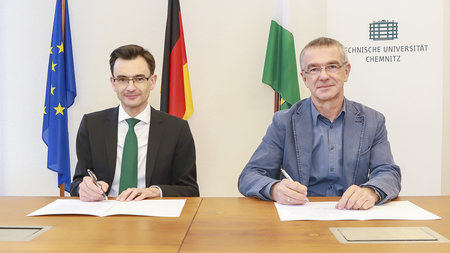 Gerd Strohmeier und Thomas Raschke sitzen an einem Tisch und unterzeichnen die Vereinbarung.