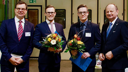 Ein Bild von vier Männern im Anzug, auf welchem zwei Personen in der Mitte einen Blumenstrauß halten