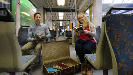 Martin Bauschmann und Carolin Ahnert sitzen in einer Straßenbahn und halten Bücher in ihren Händen. 