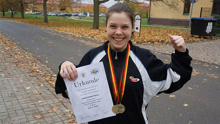 Junge Frau mit Goldmedaille lacht und hält eine Urkunde.