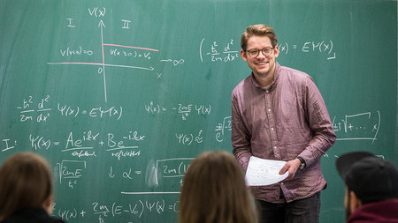 Junger Mann mit Brille steht vor einer Tafel.