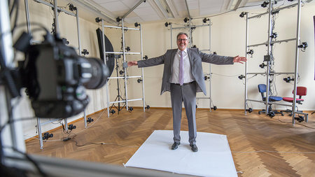 Mann mittleren Alters mit Brille steht in einem Raum. Um ihn sind Kameras.