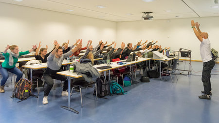 Studierende, die an Tischen sitzen, wiederholen die Bewegungsübungen, die ein Mann vor ihnen demonstriert.