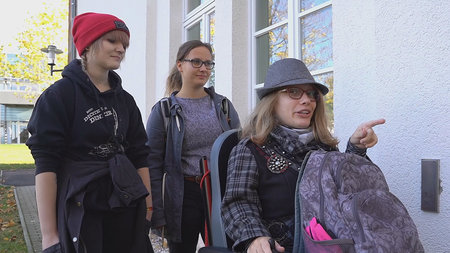 Drei junge Frauen, eine im Rollstuhl, stehen vor einer Tür mit einem elektronischen Türöffner.