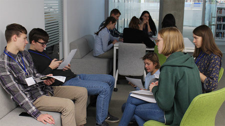 Junge Menschen sitzen in einer Bibliothek an Tischen und sprechen miteinander.