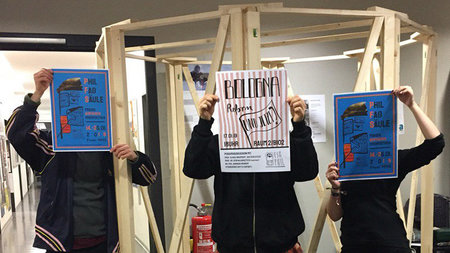 Drei Personen halten vor einem Holzgestell stehend Plakate hoch