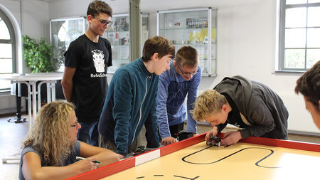 Vier Jungen und eine Frau stehen an einem Tisch, darauf stellt ein Junge einen vierrädrigen Miniroboter.