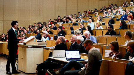 Mann steht am Rednerpult und spricht ins gut gefüllte Auditorium 