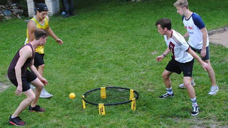 Vier Männer stehen um einen Ring, der mit einem Netz bespannt ist und versuchen einen gelben Ball zu treffen.