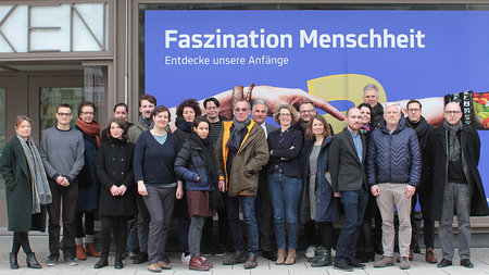 Männer und Frauen stehen vor einem Plakat mit der Aufschrift "Faszination Menschheit".