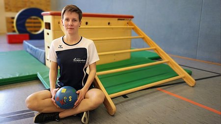 Junger Frau seitzt in einer Sporthalle mit einem Ball auf dem Schoß.