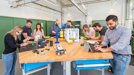 Mehrere Personen stehen an einem großen Tisch und arbeiten an verschiedenen Arbeitsplätzen mit Lötkolben, PC und anderen Geräten.