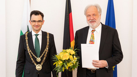 Ein Mann mittleren Alters und ein alter Mann mit Bart stehen nebeneinander. Der Ältere hält eine Urkunde und Blumen.