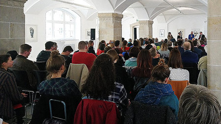 Viele Personen sitzen auf Stühlen und blicken zum Podium, wo vier Personen sitzen.