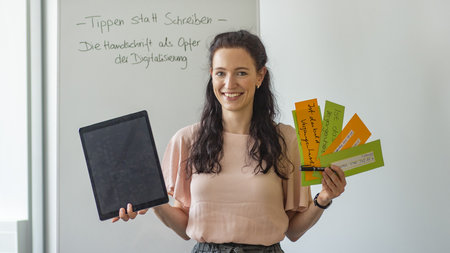 Frau steht mit Zetteln,Stift und Tablet in den Händen vor einer weißen Tafel