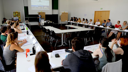 Frau steht an der Tafel, mehrere Personen sitzen an Tischen.