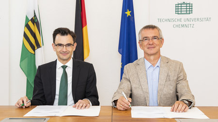 Zwei Männer mit Brille sitzen an einem Tisch und halten einen Stift in der Hand. 