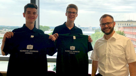 Mann steht neben zwei Jungen die beide ein T-Shirt mit dem Schriftzug Technische Universität Chemnitz hochhalten