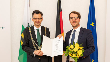 Ein Mann mittleren Alters und ein junger Mann mit Brille stehen nebeneinander und halten eine Urkunde.