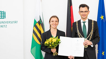 Ein Mann mittleren Alters und eine jüngere Frau stehen nebeneinander und halten eine Urkunde.