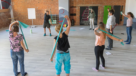 7 Personen stehen im Kreis und halten ein Gymnastikband hinter dem Rücken.