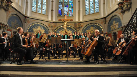 Mehrere Musiker sitzen auf Stühlen mit Instrumentenin der Hand, dahinter stehen die Chormitglieder